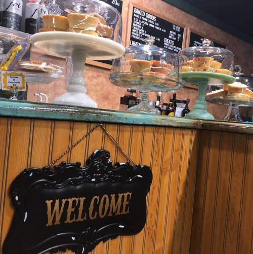 A cozy bakery display with cakes on pedestals and a "WELCOME" sign hanging on the wooden counter.