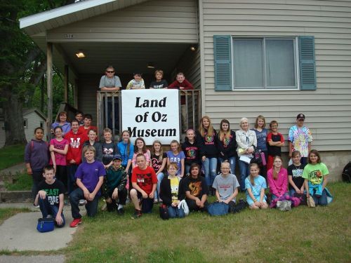 A large group of children and adults poses in front of the Land of Oz Museum, smiling and holding a sign.