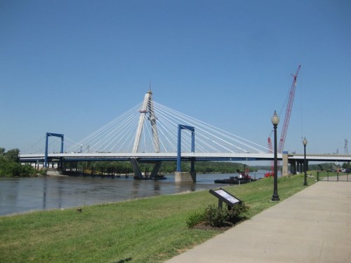 A cable-stayed bridge spans a river, with cranes in the background and a grassy area in the foreground under a clear blue sky.