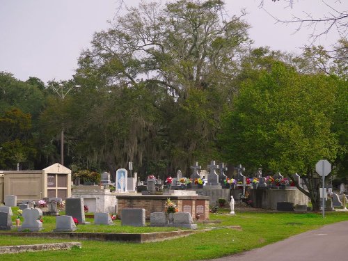 A peaceful cemetery with gravestones, crosses, and colorful flowers, surrounded by trees and a quiet road.