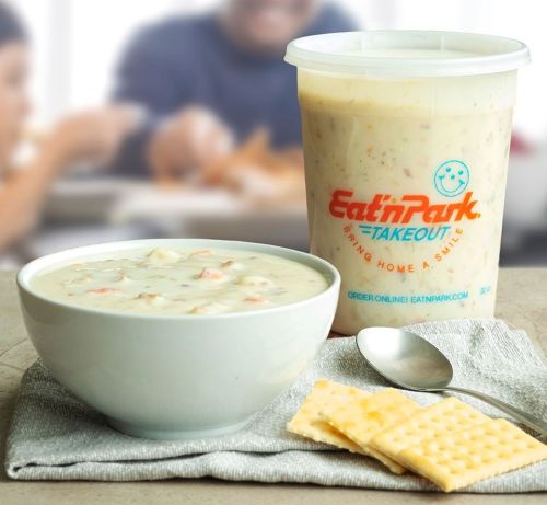 A bowl of creamy soup next to a container of Eat'n Park takeout, with crackers on a napkin in the foreground.