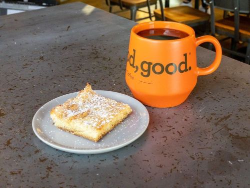 A slice of dessert dusted with powdered sugar on a plate next to a large orange coffee mug.