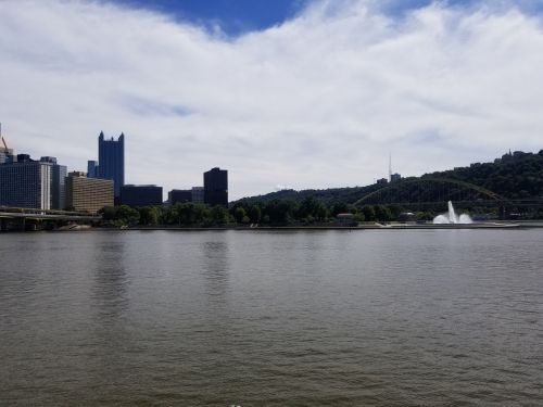 City skyline of Pittsburgh with a river in the foreground, featuring buildings, a bridge, and a fountain.
