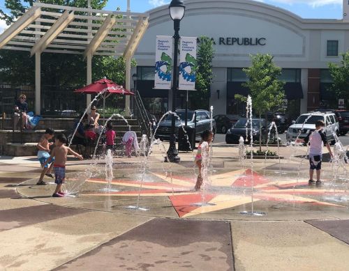 Children play in a splash pad, running through water jets on a sunny day, with shops in the background.