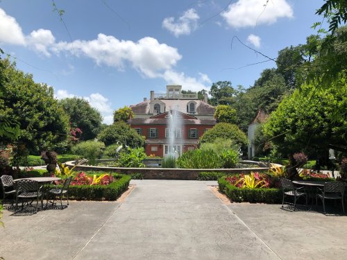 A scenic garden with a fountain, colorful plants, and a large red building in the background under a blue sky.