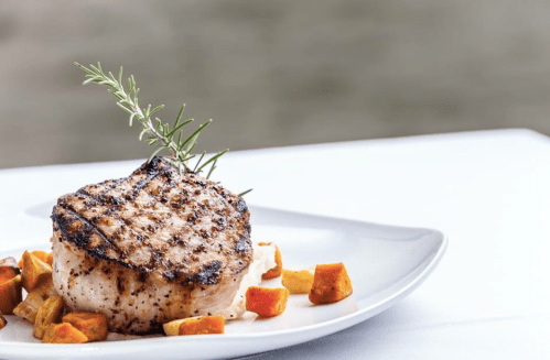 Grilled pork chop garnished with rosemary, served with roasted vegetables on a white plate.