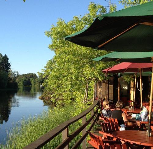 A serene lakeside scene with people sitting at wooden tables under umbrellas, surrounded by greenery and calm water.