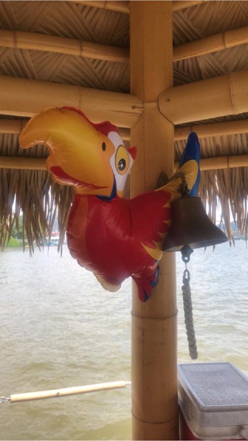 A colorful inflatable parrot hangs next to a bell on a bamboo structure by the water.