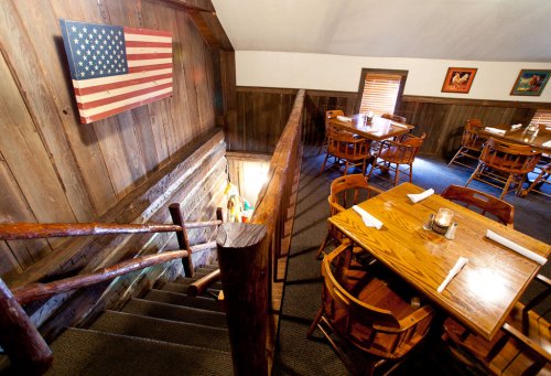 A rustic restaurant interior with wooden decor, tables, and an American flag on the wall, viewed from a staircase.