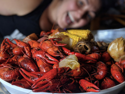 A close-up of a large pile of red crawfish and corn, with a smiling person blurred in the background.