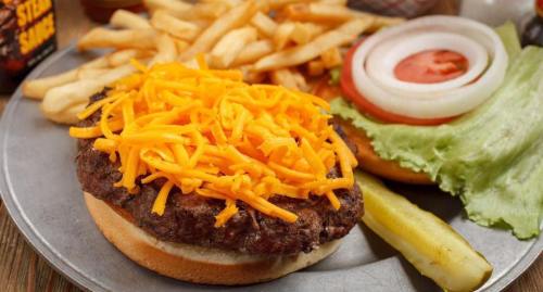 A plate with a cheeseburger topped with shredded cheese, lettuce, tomato, onion, fries, and a pickle on the side.