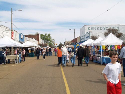 A bustling street market with vendors under tents, people browsing, and colorful displays along a wide road.