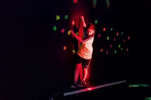 A child climbs a brightly colored rock wall in a dimly lit room, showcasing determination and focus.