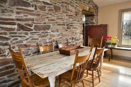 A rustic dining area with a stone wall, wooden table, and chairs, featuring a flower vase on the windowsill.