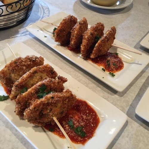 Two plates of crispy fried dumplings on skewers, served with a red sauce and garnished with parsley.