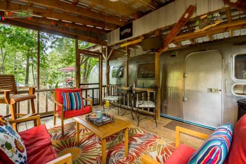 Cozy outdoor seating area with colorful cushions, a wooden table, and an Airstream trailer in a wooded setting.
