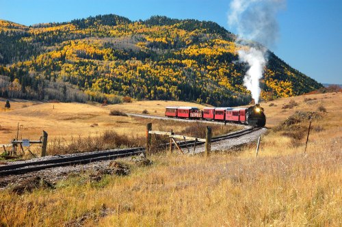 A vintage steam train travels through a scenic landscape with autumn-colored trees and rolling hills.