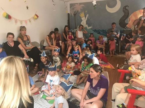 A group of children and adults sit together in a colorful room, many wearing paper crowns, engaged in an activity.