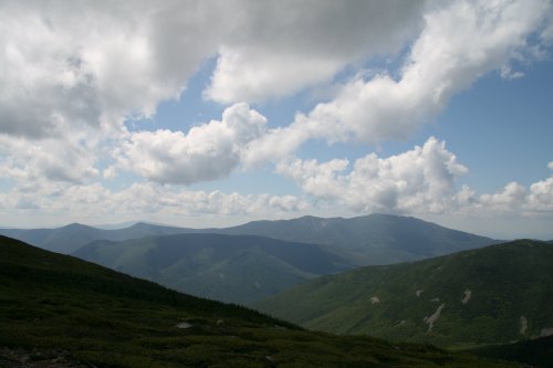 A panoramic view of green mountains under a partly cloudy sky, showcasing rolling hills and distant peaks.