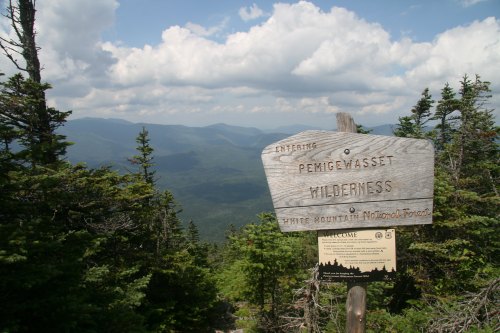 A wooden sign marks the entrance to Pemigewasset Wilderness, with mountains and trees in the background under a cloudy sky.