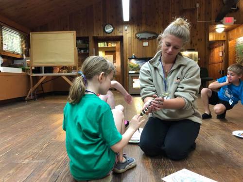 A woman kneels beside a girl, showing her a small animal in a wooden room with children engaged in activities.