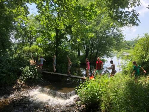 A group of people walking on a wooden bridge over a small waterfall, surrounded by lush greenery and a river.