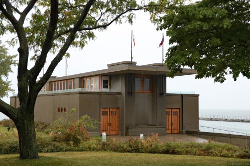 A modernist building with a flat roof, surrounded by greenery and near a body of water, under a cloudy sky.