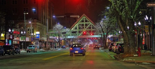 A nighttime street scene with a blue car driving, illuminated buildings, and a distinctive glass-roofed structure overhead.