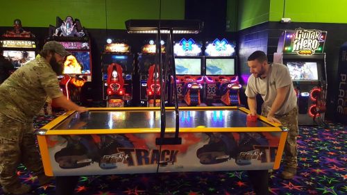 Two men play air hockey in a colorful arcade filled with various games and bright lights.