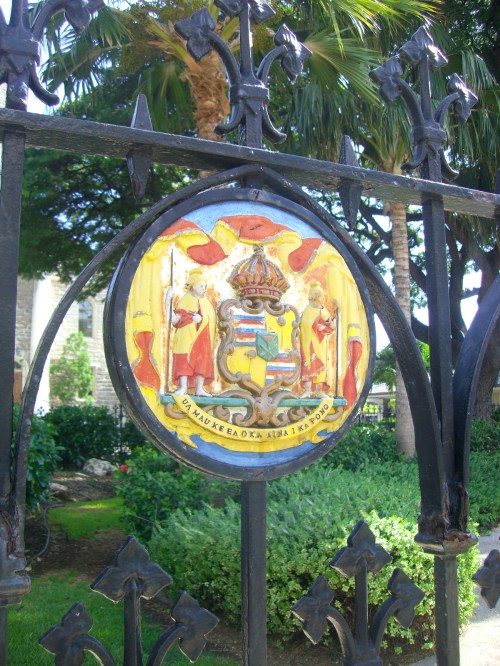 Colorful emblem on a wrought iron gate, featuring a crown and figures in red robes, surrounded by greenery.