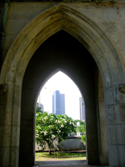 A stone archway frames a view of modern buildings and greenery in the background.