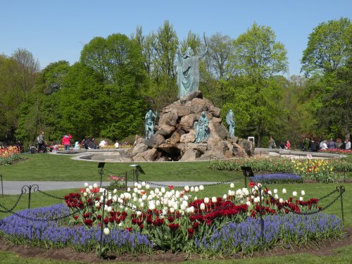 A scenic park with a statue on a rock, surrounded by colorful flower beds and lush green trees under a clear blue sky.