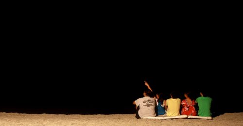 A group of four people sitting on a beach at night, looking up at the stars, with one pointing towards the sky.
