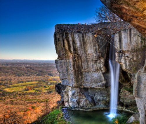 A stunning waterfall cascades from a rocky cliff into a serene pool, surrounded by vibrant autumn foliage and distant mountains.