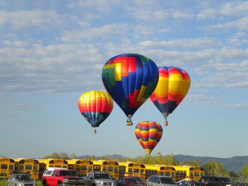 Colorful hot air balloons float in a clear sky above parked yellow school buses.