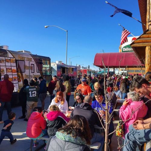 A bustling food truck event with a large crowd enjoying food under a clear blue sky. Flags and food trucks in the background.
