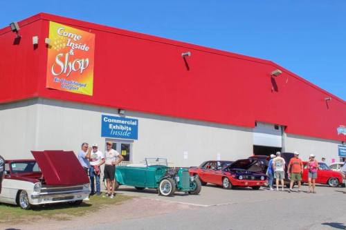 A group of people stands outside a red building with classic cars parked in front during a car show event.