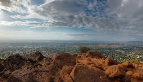 A panoramic view from a rocky summit, overlooking a sprawling cityscape under a cloudy sky.