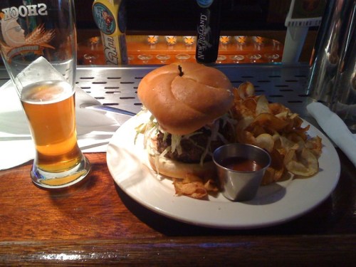A plate with a burger topped with lettuce, served with a side of chips and a glass of beer on a wooden bar.