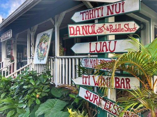 Colorful signs point to various attractions outside a tropical-themed shop, surrounded by lush greenery.