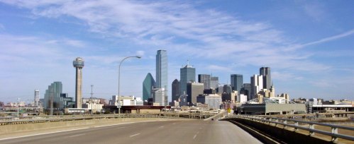 View of a city skyline with modern skyscrapers and a clear blue sky, seen from an empty highway.