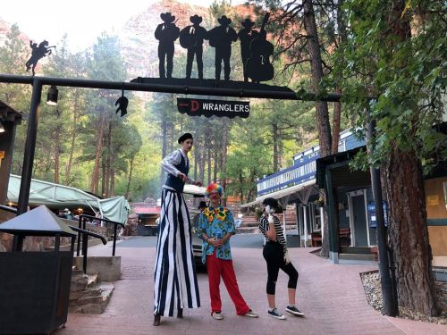A stilt walker and a clown pose in a scenic outdoor area with trees and mountains in the background.