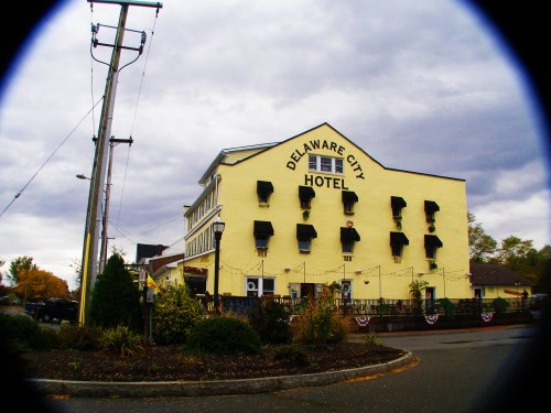 A yellow hotel building with black shutters, surrounded by greenery and a cloudy sky, labeled "Delaware City Hotel."
