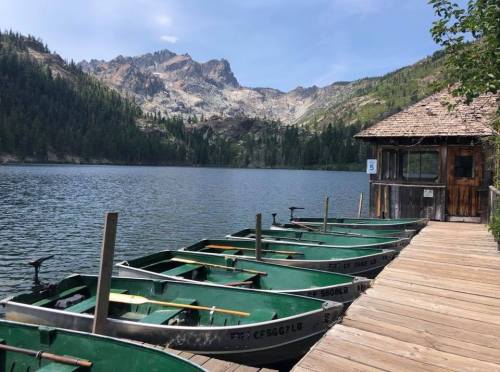 A serene lake with green boats docked at a wooden pier, surrounded by mountains and lush trees.
