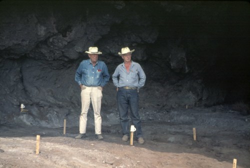 Two men in hats stand in a cave, with excavation markers visible in the foreground.