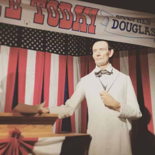 A lifelike wax figure of Stephen Douglas stands at a podium, with a patriotic backdrop of flags and a banner.