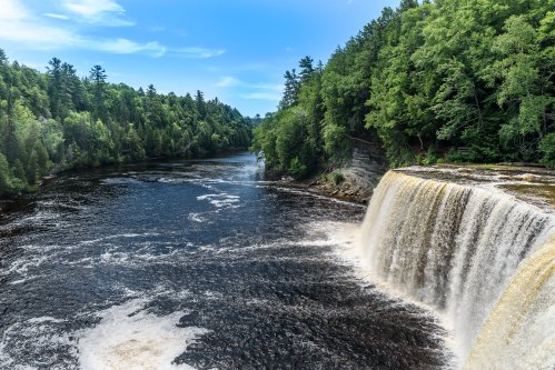 A scenic view of a waterfall cascading into a river, surrounded by lush green trees under a clear blue sky.
