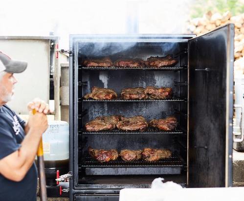 A man stands beside a smoker filled with racks of sizzling meat, with smoke rising from the top.