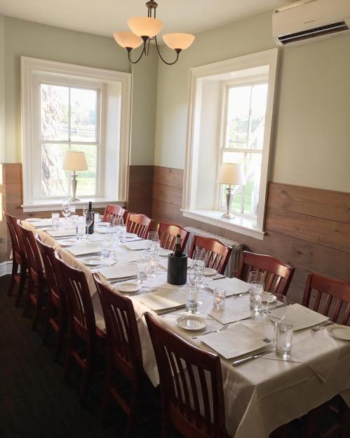 A dining room with a long table set for a meal, surrounded by wooden chairs and natural light from windows.