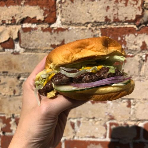 A hand holding a juicy burger with lettuce, onion, pickles, and cheese against a brick wall background.